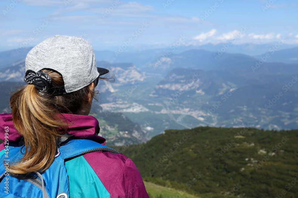 The girl standing backwards with the backpack looks at the Adige valley from the Paganella peak in Trentino.