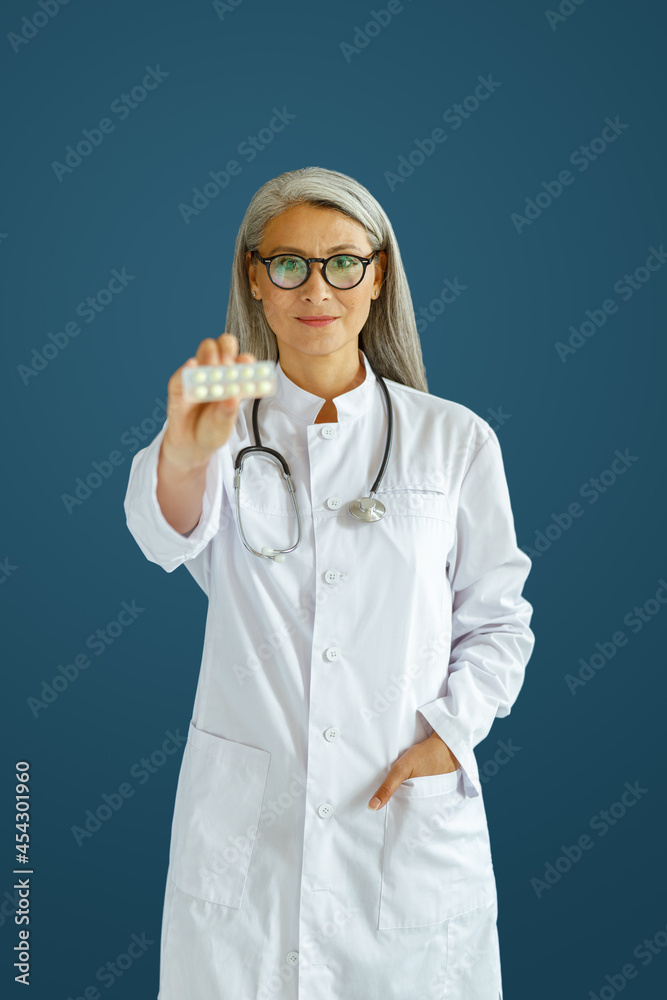 Cheerful mature woman doctor in white robe shows pills blister posing on blue background in studio. Professional medical staff Cheerful mature woman doctor in white robe shows pills blister posing on blue background in studio. Professional medical staff