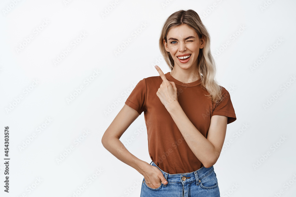 Smiling blond girl winking at you, pointing finger at upper left corner sale advertisement, showing smth, standing against white background Smiling blond girl winking at you, pointing finger at upper left corner sale advertisement, showing smth, standing against white background