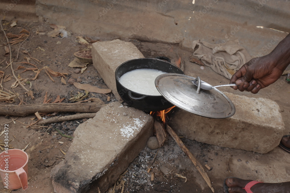 horizontal photography of a black metal pot with rice boiling inside, standing on three concrete bricks over burning wood fire, outdoors in the Gambia, Africa