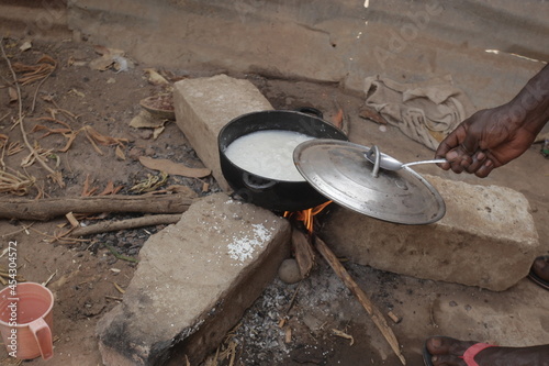 horizontal photography of a black metal pot with rice boiling inside  standing on three concrete bricks over burning wood fire  outdoors in the Gambia  Africa