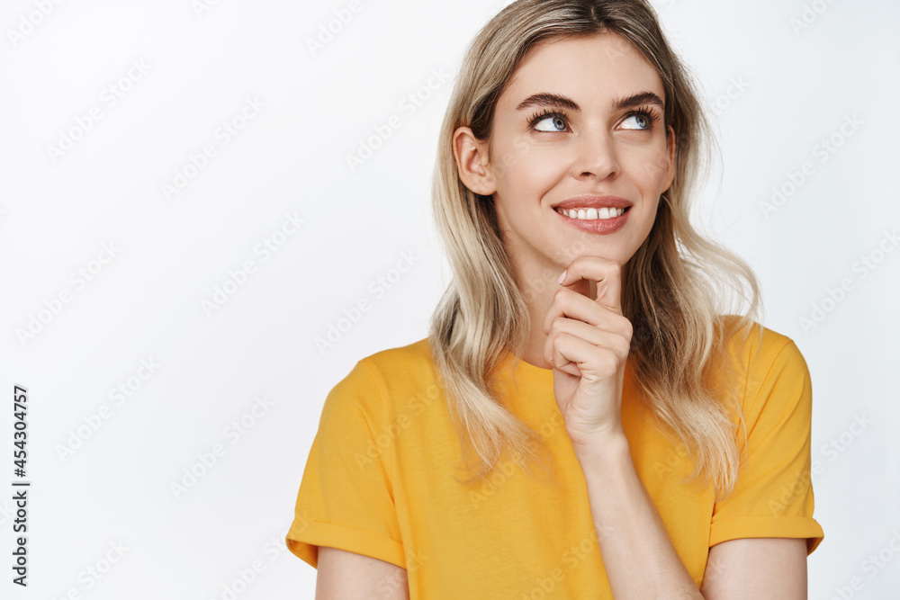 Young blond woman smiling, thinking and looking aside with satisfied face, has an idea, standing in yellow t-shirt against white background Young blond woman smiling, thinking and looking aside with satisfied face, has an idea, standing in yellow t-shirt against white background
