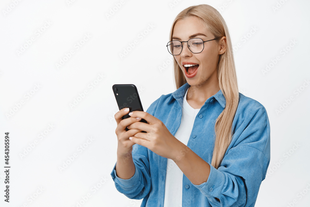 Cellular and smartphone technology. Young cute girl in glasses looking at her mobile phone with surprised face, standing over white background Cellular and smartphone technology. Young cute girl in glasses looking at her mobile phone with surprised face, standing over white background