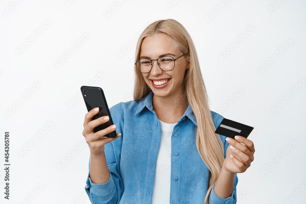 Happy blond woman smiling, wearing glasses, looking at mobile phone screen while paying for online order, holding credit card, standing over white background Happy blond woman smiling, wearing glasses, looking at mobile phone screen while paying for online order, holding credit card, standing over white background