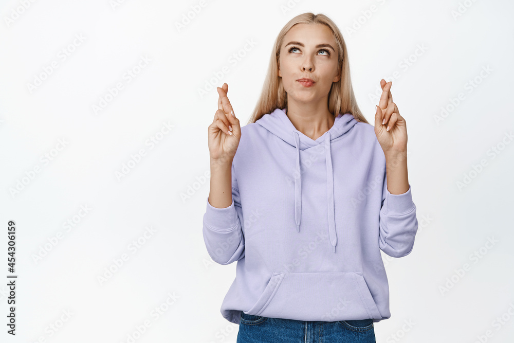 Hopeful blond girl cross fingers for good luck and looking at upper left corner, praying, making wish for dream come true, standing over white background Hopeful blond girl cross fingers for good luck and looking at upper left corner, praying, making wish for dream come true, standing over white background