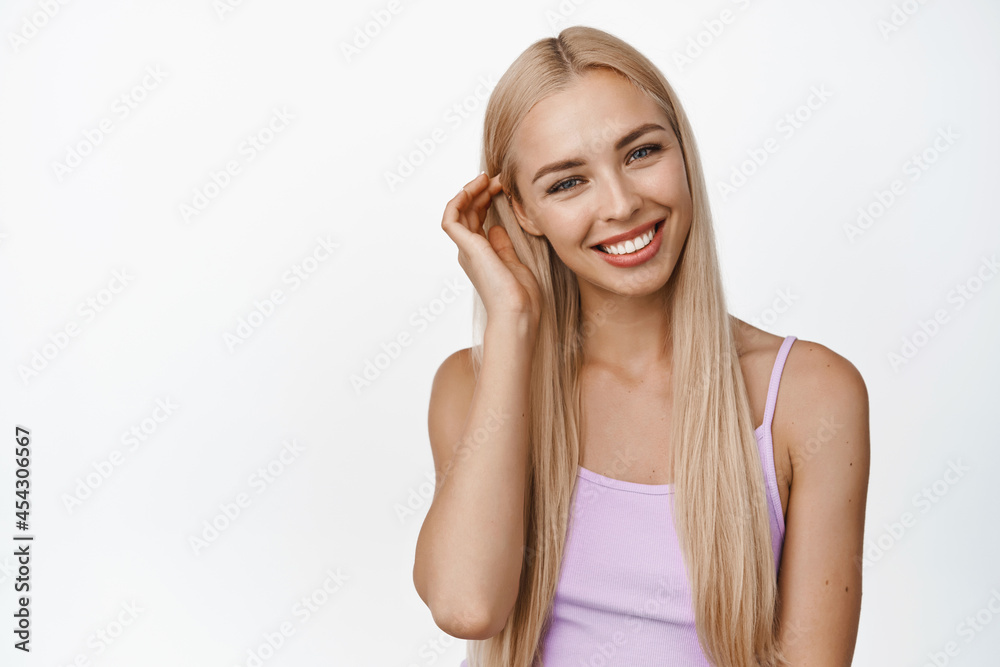 Cute smiling girl tuck hair behind ear and looking happy at camera, concept of women skin care and wellbeing, standing against white background Cute smiling girl tuck hair behind ear and looking happy at camera, concept of women skin care and wellbeing, standing against white background