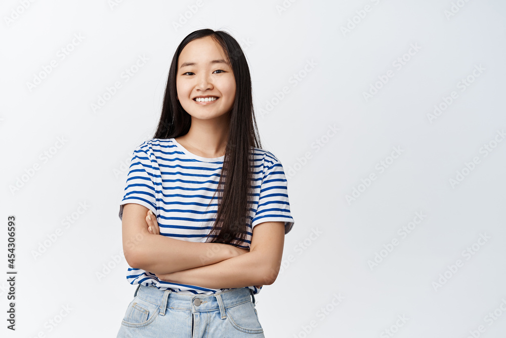 Smiling asian girl looking happy, cross arms on chest, standing in striped t-shirt over white background Smiling asian girl looking happy, cross arms on chest, standing in striped t-shirt over white background
