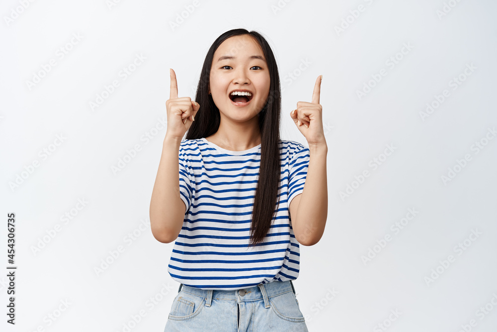 Excited asian girl pointing fingers up and laughing, smiling while showing advertisement, inviting clients to shop, recommending store, standing over white background Excited asian girl pointing fingers up and laughing, smiling while showing advertisement, inviting clients to shop, recommending store, standing over white background