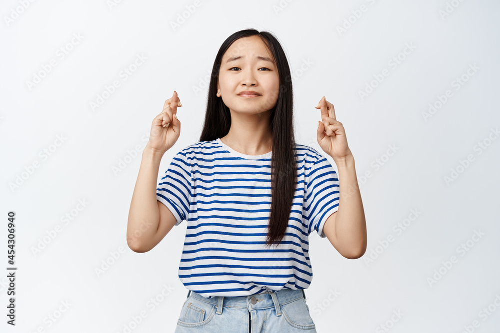 Nervous asian girl looking hopeful at camera, praying with fingers crossed, begging or anticipating, standing in tshirt against white background Nervous asian girl looking hopeful at camera, praying with fingers crossed, begging or anticipating, standing in tshirt against white background