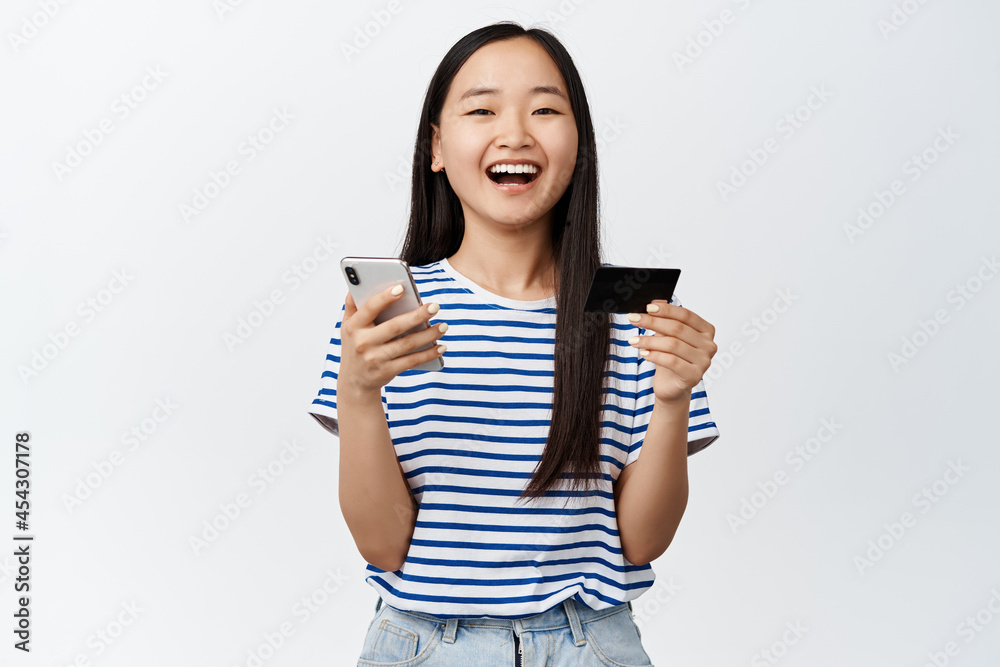 Online shopping and finance app. Smiling asian girl laughing, holding smartphone with credit card, paying in application, standing over white background Online shopping and finance app. Smiling asian girl laughing, holding smartphone with credit card, paying in application, standing over white background