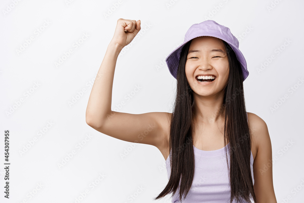 Cheerful asian girl in bucket hat celebrating, raising fist up for support, being proactive, chanting and smiling, standing over white background Cheerful asian girl in bucket hat celebrating, raising fist up for support, being proactive, chanting and smiling, standing over white background