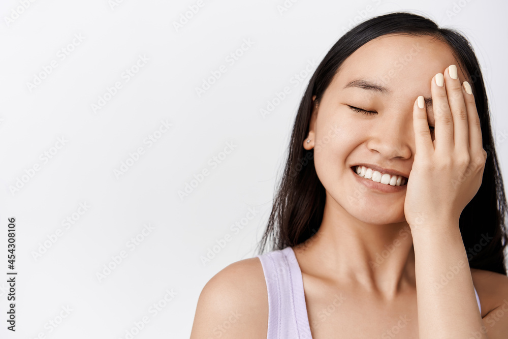 Skin care. Happy asian woman touching face with pleased smiling face, close eyes, using cosmetic product on facial skin, standing against white background Skin care. Happy asian woman touching face with pleased smiling face, close eyes, using cosmetic product on facial skin, standing against white background