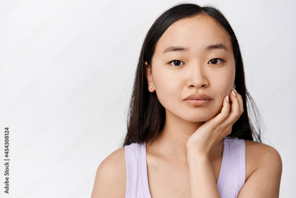 Close up portrait of asian girl touching face and gazing at camera, promo of skin care cosmetic product, cleanser or moisturizer, standing over white background Close up portrait of asian girl touching face and gazing at camera, promo of skin care cosmetic product, cleanser or moisturizer, standing over white background