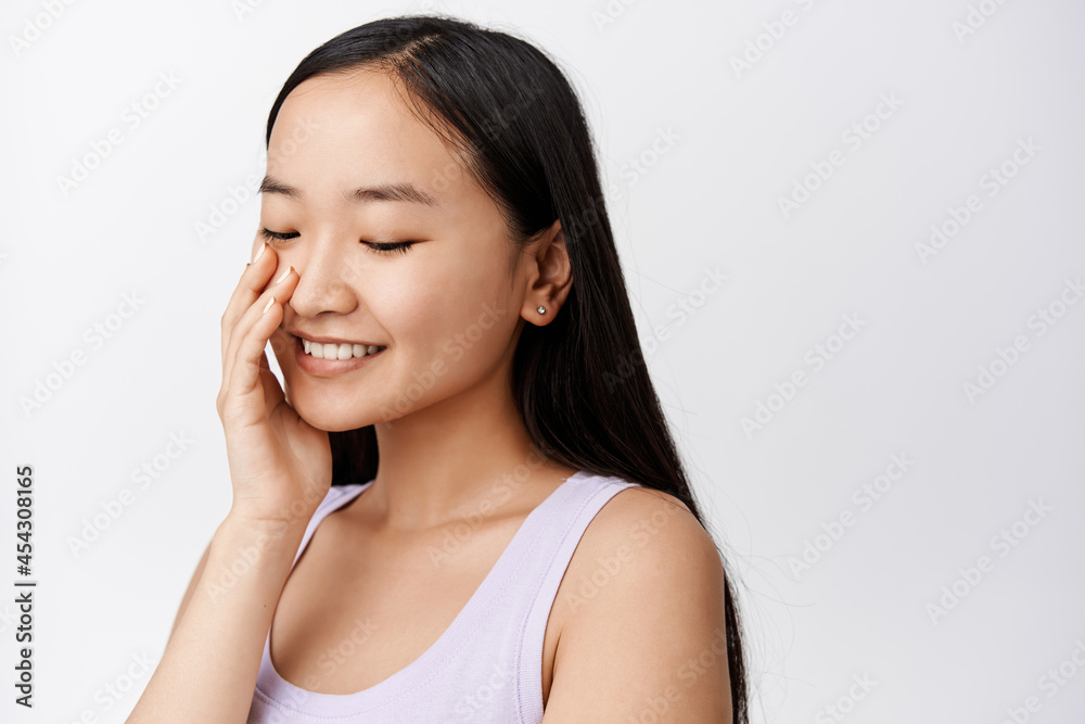 Tender asian girl gently touching soft facial skin after using skincare moisturizer, cleansing cosmetic product, smiling happy, white background Tender asian girl gently touching soft facial skin after using skincare moisturizer, cleansing cosmetic product, smiling happy, white background
