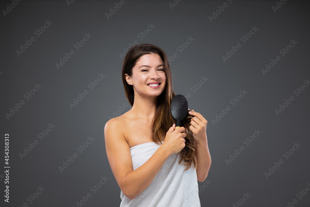 Beautiful and well-groomed woman. Female wrapped in a white towel around her body stands in front of a gray wall and uses a hair brush after showering. Natural beauty and femininity Hair straightening Beautiful and well-groomed woman. Female wrapped in a white towel around her body stands in front of a gray wall and uses a hair brush after showering. Natural beauty and femininity Hair straightening