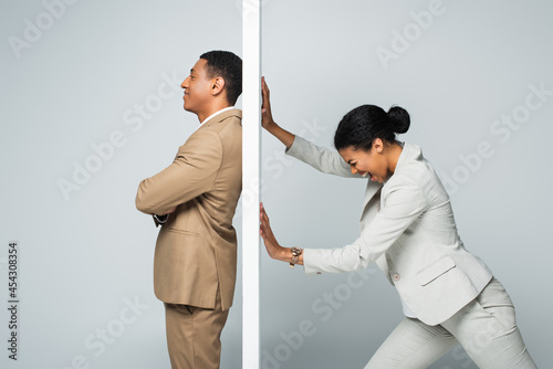 angry african american businesswoman screaming and pulling wall near smiling businessman on grey photo
