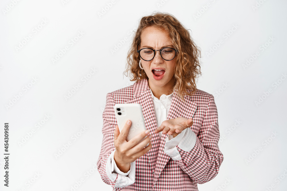 Angry corporate woman, lady boss looking at mobile phone and screaming frustrated, standing bothered against white background Angry corporate woman, lady boss looking at mobile phone and screaming frustrated, standing bothered against white background