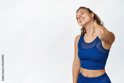 Image of satisfied sports girl rub her neck and smile. Fitness woman feeling pleasure after doing productive workout, doing exercises, white background