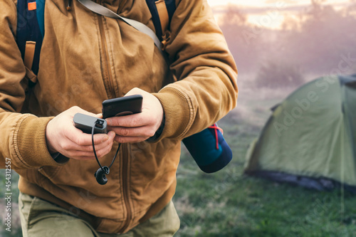 Fototapeta Naklejka Na Ścianę i Meble -  Man holds a smartphone in his hands and charges it with a power bank against the backdrop of a tourist tent in nature. Portable travel charger.