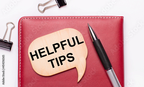 On a light background there are black paper clips, a pen, a burgundy notepad a wooden board with the text HELPFUL TIPS