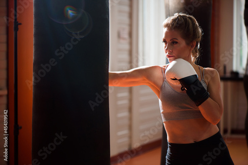 Young woman doing boxing workout in the gym, she is wearing boxing gloves and punching a punching bag. © nagaets