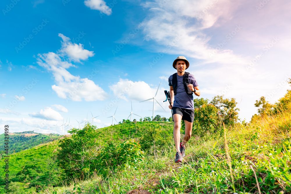 Asian male trail runners, wearing sportswear, are practicing running on a high mountain behind a beautiful view. There is a field of wind turbines generating electricity in the background.