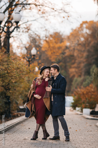 A couple of beautiful lovers spend time outdoors on the fall day. Pretty woman in a red dress and modish hat and a handsome man in a raincoat posing at the city park on a sunny autumn day © innarevyako