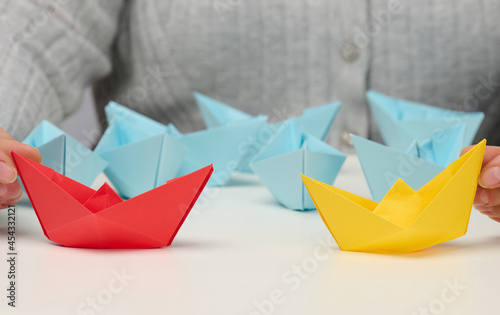 female hands hold red and yellow paper boats on a white table. Confrontation, follow a strong leader, dailog photo