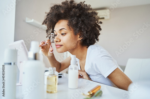 Focused young woman using an eyelash curler at home