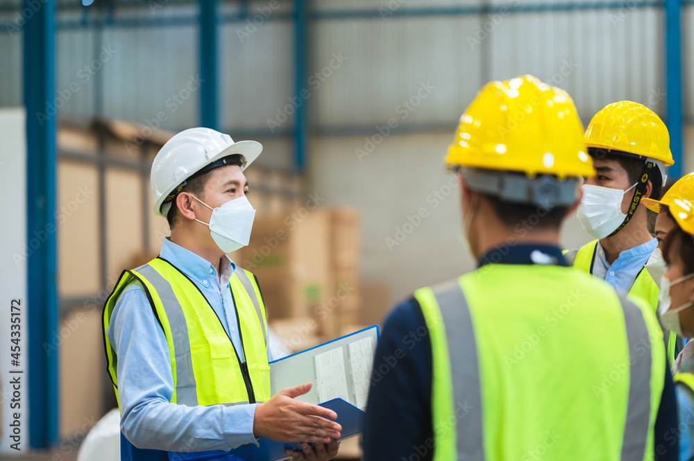The factory employee consists of engineers, foreman, technicians, and related department staff. Wear a mask, hard hat, and vest. meeting before starting work inside the warehouse. Teamwork concept. The factory employee consists of engineers, foreman, technicians, and related department staff. Wear a mask, hard hat, and vest. meeting before starting work inside the warehouse. Teamwork concept.