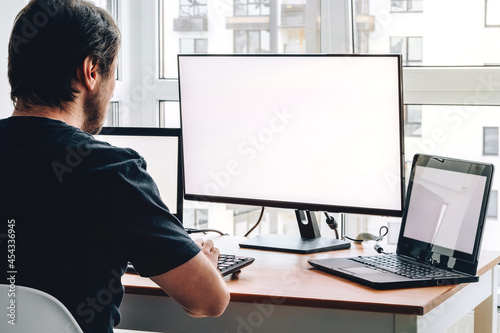 Obraz na plátně Man sitting at workplace with two laptops and monitor near the window