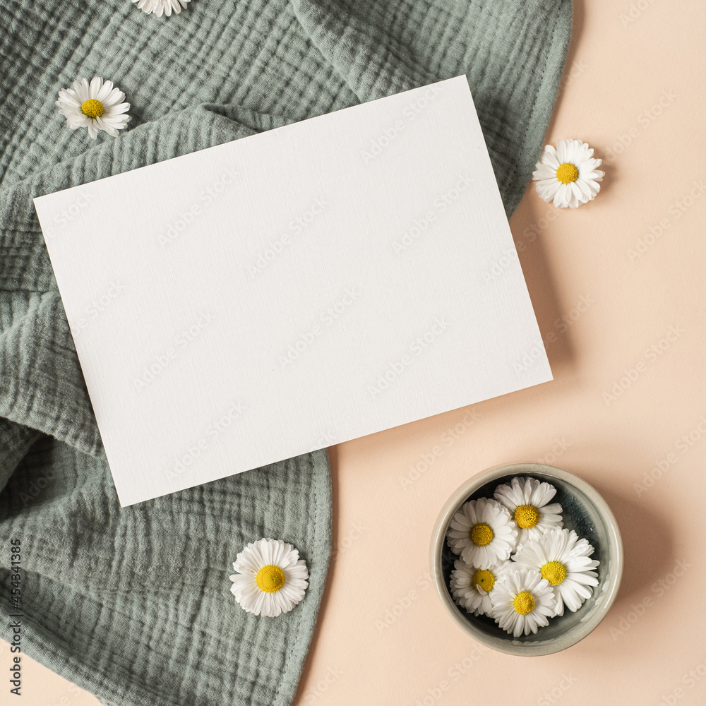 Obraz premium Flatlay aesthetic Parisian styled invitation template. Blank paper sheet card with mockup copy space, chamomile flower buds and aquamarine muslin cloth on neutral beige background. Flat lay, top view