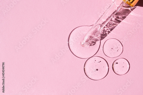 Transparent drops of hyaluronic acid and glass pipette on pink background.  Glass pipette with a drop of face serum with air bubbles. Top view, place for text.  photo