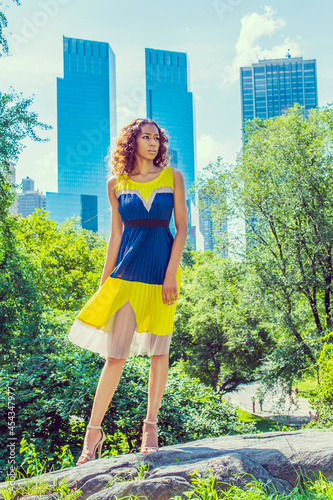 Dressing in a sleeveless, yellow and blue pattern dress, a young pretty black college student with long curly hair is confidently standing in the front of a business district..