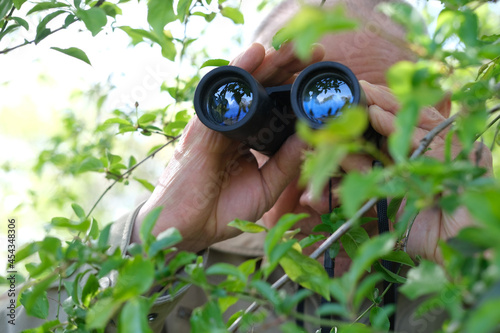 adult man holding black field binoculars with zoom, hiding in greenery, peeping out of green bushes, hunts down secrets, concept of surveillance, observation, investigating crime, watching his wife