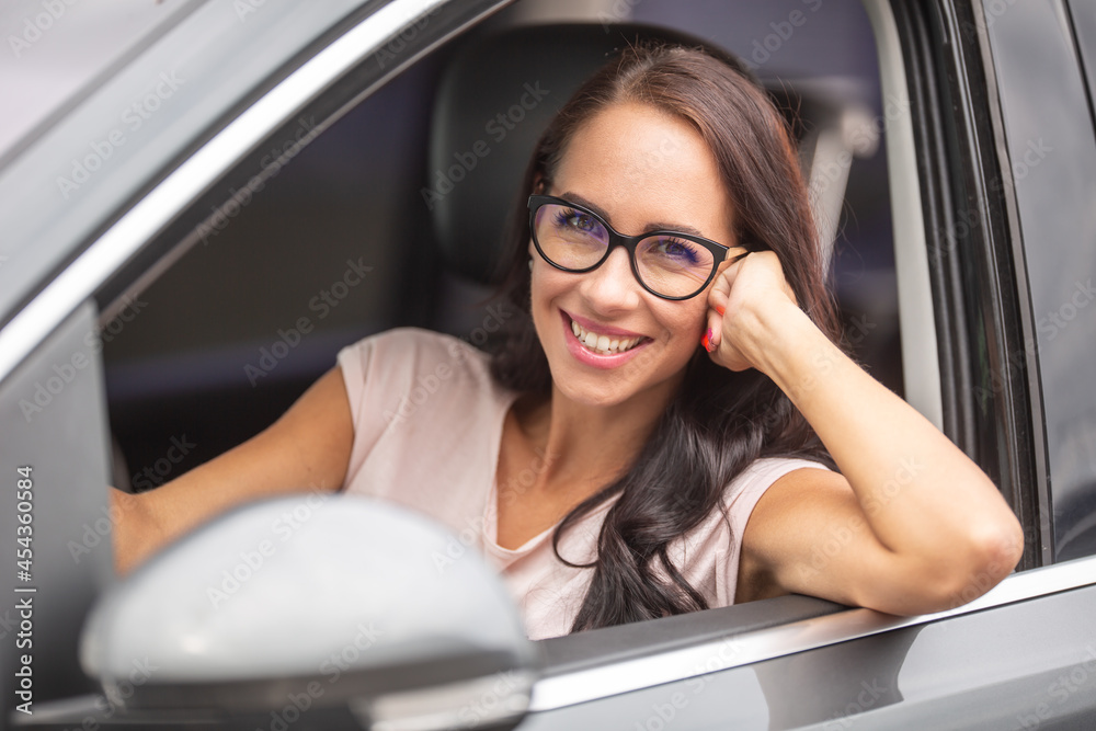 Smiling brunette with glasses drives a car, with one arm out of the open window on the side of her car