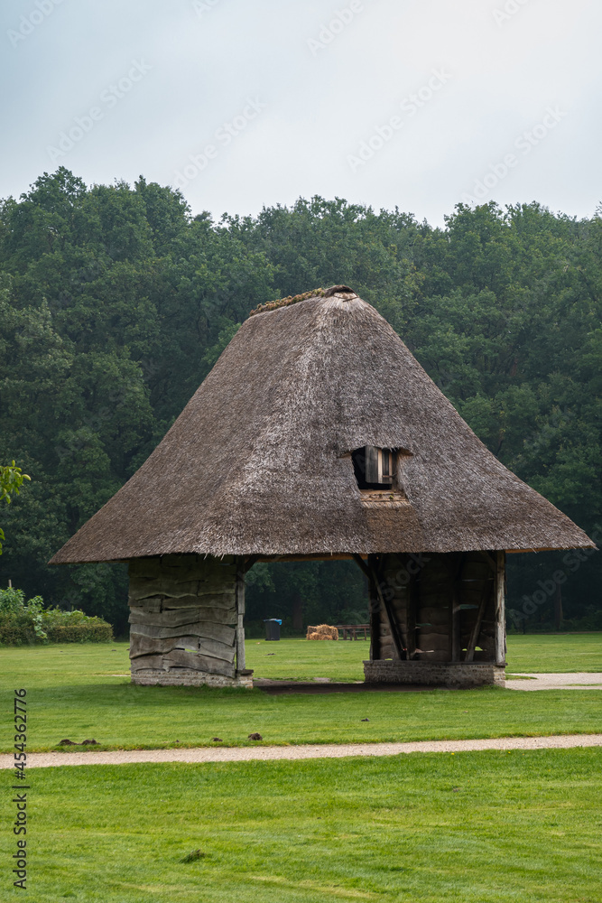 Genk, Belgium - August 11, 2021: Domein Bokrijk. Drive through barn with thick gray straw roof with small window under light blue sky, set in meadow with dark green foliage in back. Genk, Belgium - August 11, 2021: Domein Bokrijk. Drive through barn with thick gray straw roof with small window under light blue sky, set in meadow with dark green foliage in back.