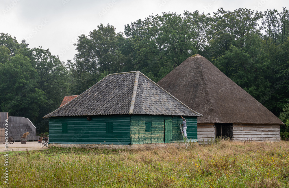 Genk, Belgium - August 11, 2021: Domein Bokrijk. 2 barns, one is painted and has tile roof, while the other. the biggest has thick straw roof. Green meadow in front, darker green foliage in back.