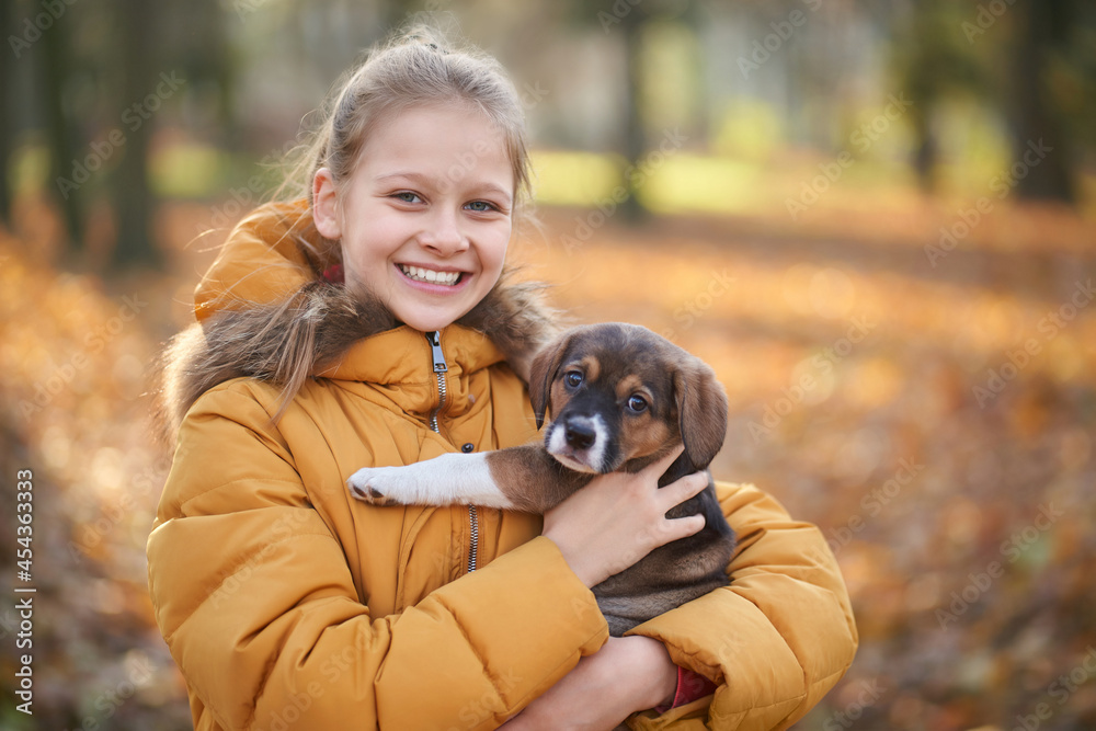 Front view of pretty girl in yellow jacket holding small dog on forest background. Concept of walking child in forest with dog. Front view of pretty girl in yellow jacket holding small dog on forest background. Concept of walking child in forest with dog.