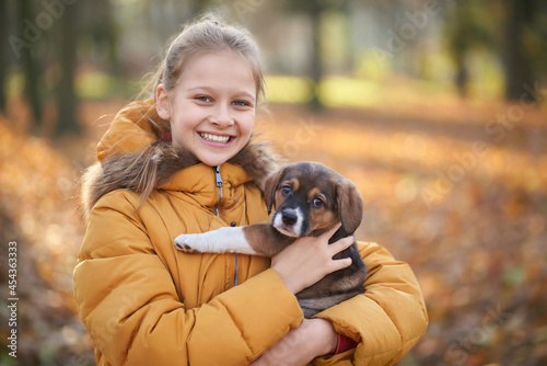 Front view of pretty girl in yellow jacket holding small dog on forest background. Concept of walking child in forest with dog.