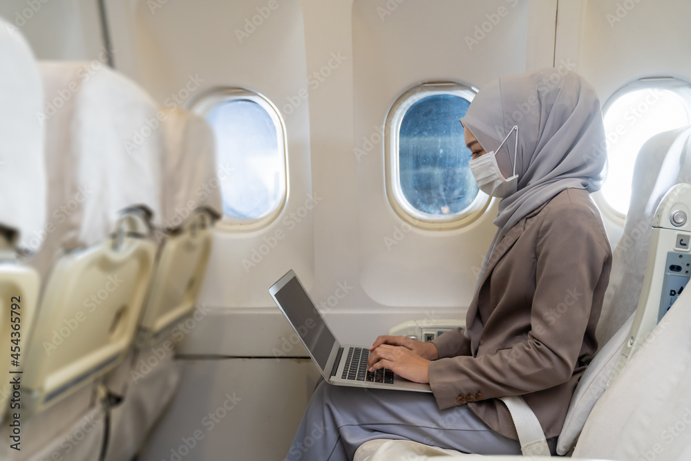 Asian Muslim business woman in hijab headscarf wearing protective face mask and using laptop computer in airplane during coronavirus disease or COVID 19 pandemic outbreak