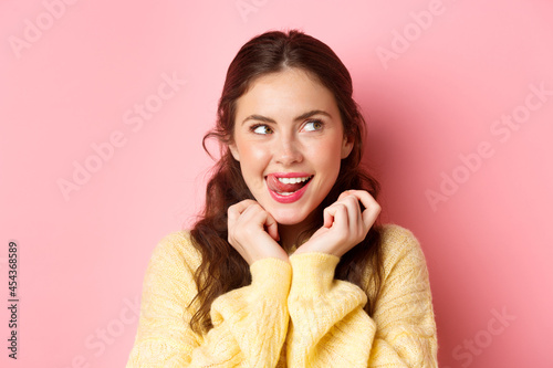 Young devious girl licking her lips and teeth, looking aside with thoughtful face, having interesting idea, thinking, standing against pink background