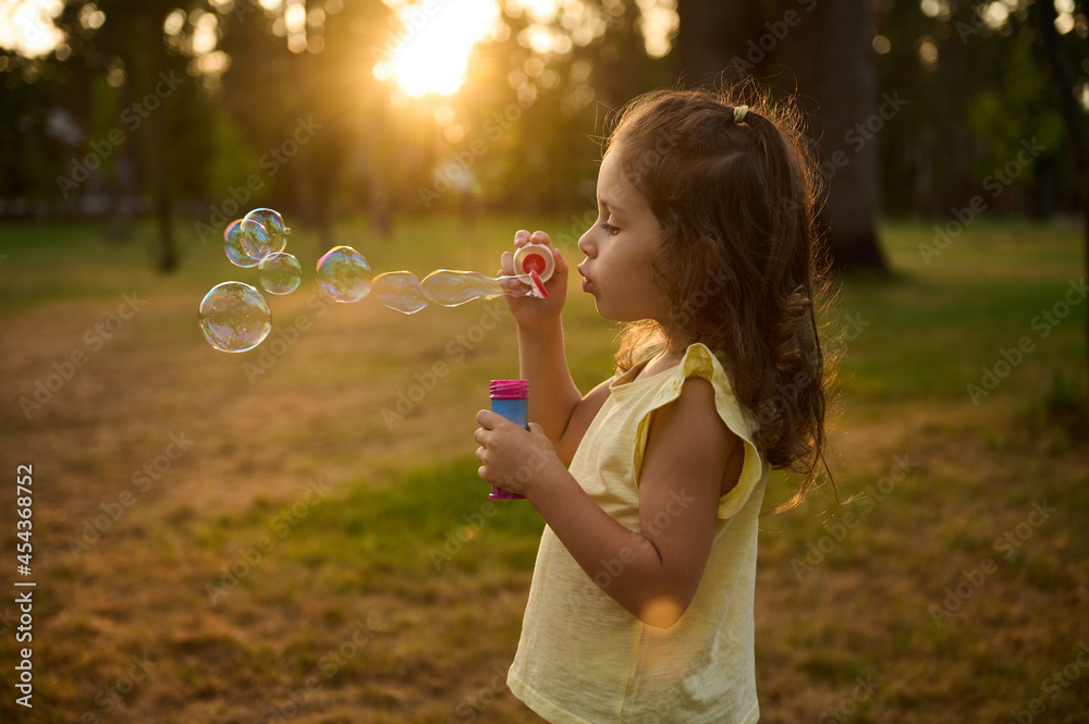 Adorable child baby girl, blowing soap bubbles at sunset, enjoying pleasant time outdoors in the meadow. Sun's rays fall through transparent bubble spheres with iridescent reflections. Adorable child baby girl, blowing soap bubbles at sunset, enjoying pleasant time outdoors in the meadow. Sun's rays fall through transparent bubble spheres with iridescent reflections.