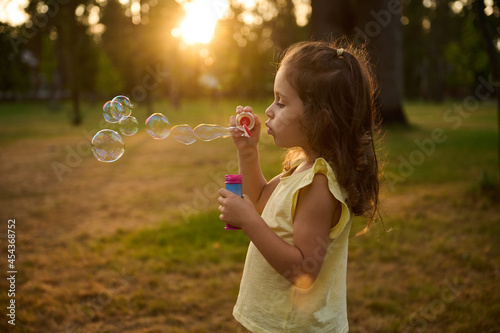 Adorable child baby girl  blowing soap bubbles at sunset  enjoying pleasant time outdoors in the meadow. Sun s rays fall through transparent bubble spheres with iridescent reflections.