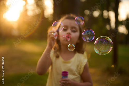 Soft focus on soap bubbles on the blurred background of a cute baby girl blowing soap bubbles in meadow, enjoying carefree childhood, recreation on the nature background at sunset