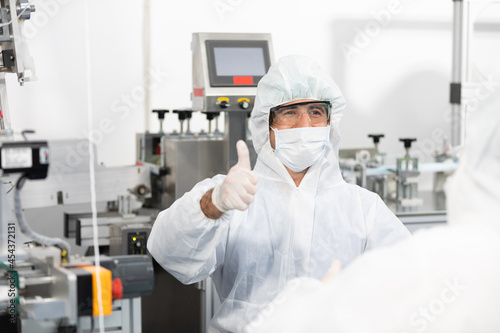male engineers wearing personal protective equipment uniform(PPE) wearing medical face mask, producing medical face masks thumbs up pose beside machine in laboratory