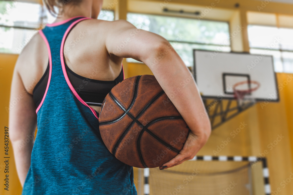 Fototapeta premium Female high school basketball player holding a ball on school gymnasium, with basketball board in background.