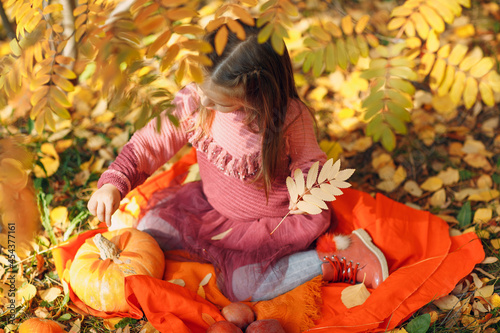 Cute little girl in autumn park with orange color leaves and yellow pumpkin