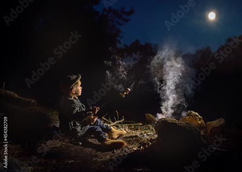 a boy is sitting by a fire in the forest and frying mushrooms on a stick