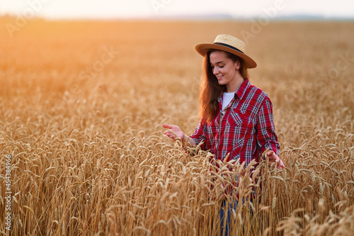 Portrait of cute young happy smiling woman farmer standing alone during walking through a yellow field of dry ripe wheat among golden spikelets at sunset
